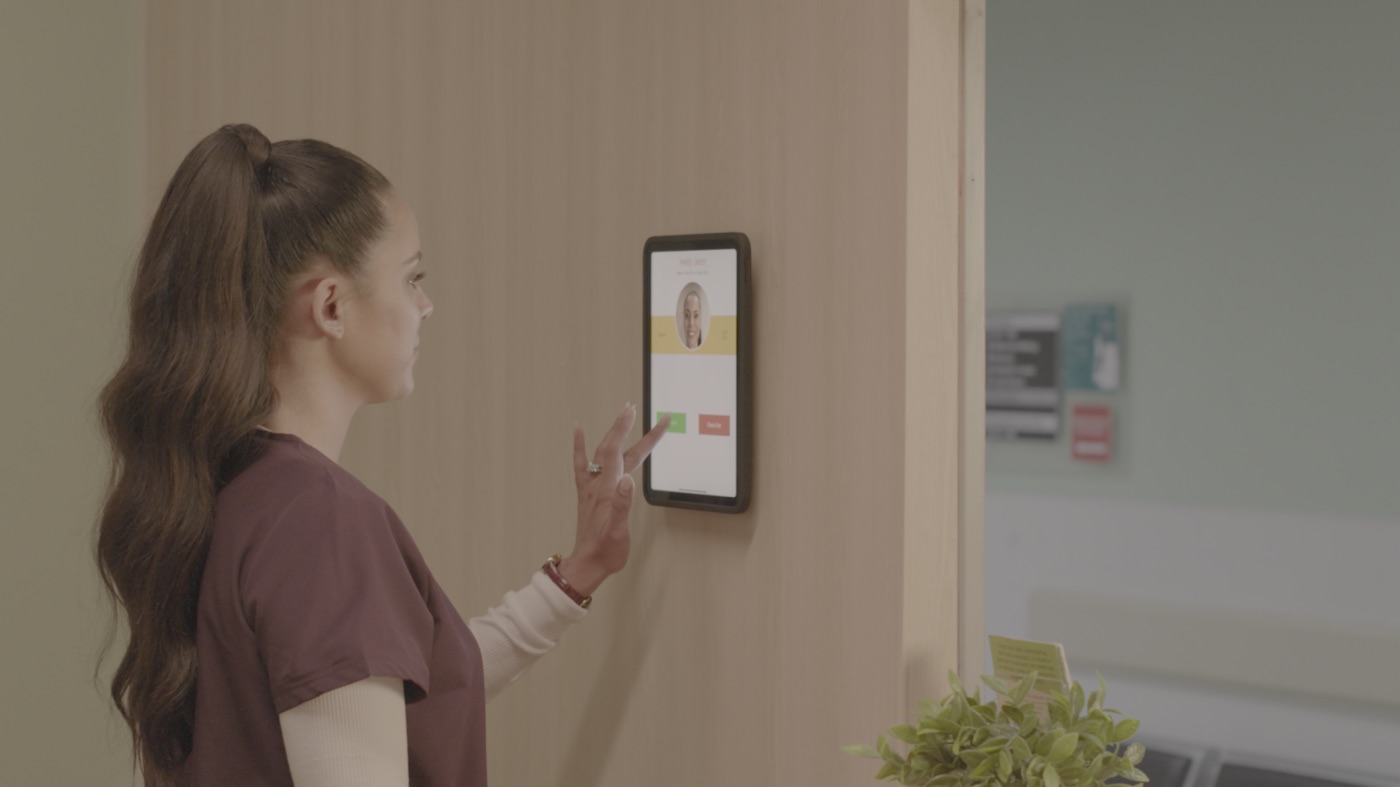 A wall-mounted iPad in an IPORT case showing the NoahFace clock-in screen in a clinic corridor